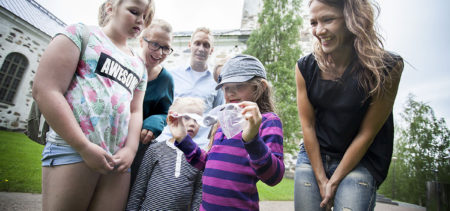 Geokätköilijät Emma Seppälä (edestä vas.), Emil Kolehmainen, Pinja ja Annette Kärki, Mari Seppälä ja Ville Kolehmainen ja lytynyt geokätköpurkki. Taustalla Kuopion tuomiokirkko.