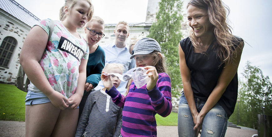 Geokätköilijät Emma Seppälä (edestä vas.), Emil Kolehmainen, Pinja ja Annette Kärki, Mari Seppälä ja Ville Kolehmainen ja lytynyt geokätköpurkki. Taustalla Kuopion tuomiokirkko.