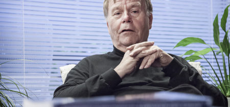 Juhani Laurinkari istuu olohuoneessa, sohvapöydällä kirjapino.
