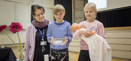 Oona Kajava pitelee Kasperi Eino Serggeiksi nimettyä vauvanukkea ja Pauli Asikainen kastemaljaa. Opettaja Anne Niemelän kanssa eikaluokkalaiset ovat tehneet kinkeritehtäviä ennen yhteistä salituntia.