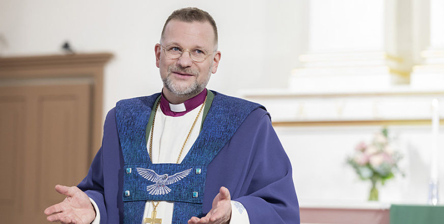 Piispa Jari Jolkkonen violetissa kirkollisessa asussaan, avoimet kädet ojennettuina.