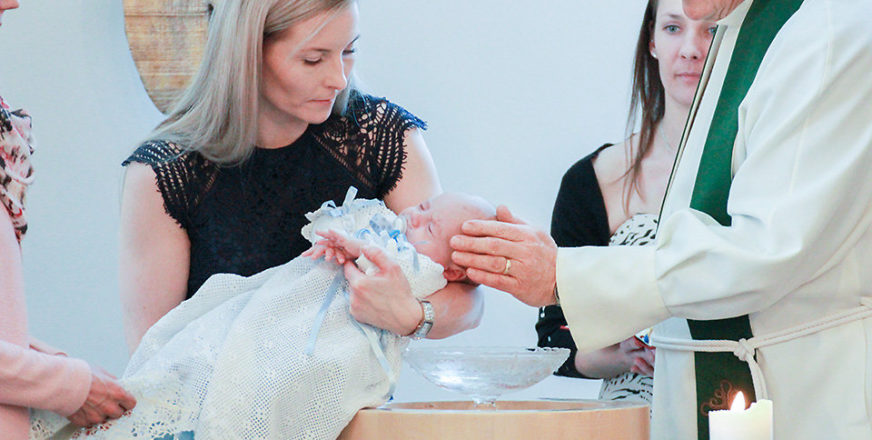 Nainen pitelee kastepukuun puettua vauvaa kastemaljan äärellä, papin käsi on lapsen pään päällä.