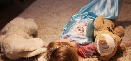 Pieni Frozen-mekkoon pukeutunut tyttö makaa lattialla selällään pehmeän maton päällä kahden pehmolelun välissä.