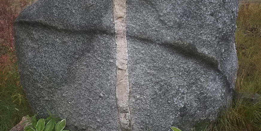 Iso harmaa kivi, johon erivärinen pystyraita ja vaakasuuntainen raita muodostavat ristin.