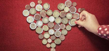 Kolikoita sydämen muotoon aseteltuna punaisella taustalla, lapsen käsi koskettaa rahaa.