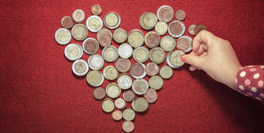Kolikoita sydämen muotoon aseteltuna punaisella taustalla, lapsen käsi koskettaa rahaa.