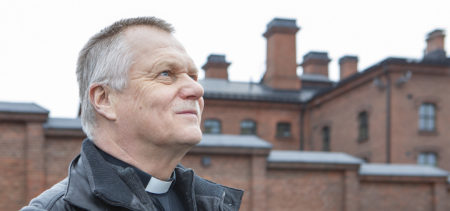 Punatiilinen vankilarakennus on taustalla, vankilapappi Jouko Koistinen katsoo lähikuvassa yläoikealle.