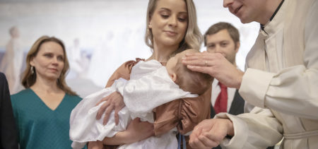 Nainen pitää kasteemekkoon puettua vauvaa sylissään, vauvan pään päällä papin käsi, taustalla kaksi aikuista.