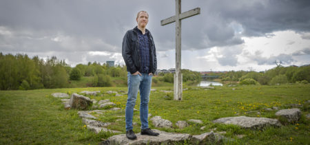 Teologi Patrik Hagman seisoo ulkona puisen ristin juurella.