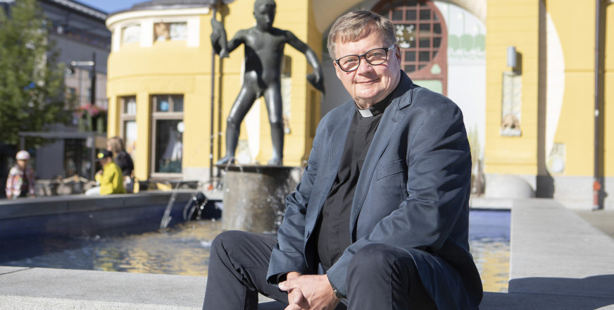 Diakoniajohtaja Seppo Marjanen istuu Veljmies-patsaan suihkulähteen äärellä, taustalla näkyy Kuopion kauppahalli.