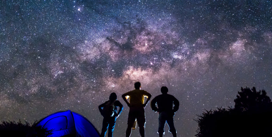 Kolme ihmistä seisoo teltan vieressä ja tähyilee tähtitaivasta.