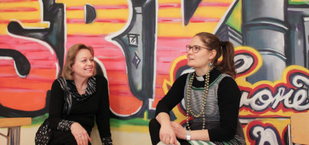 Kaksi naista istuu graffitiseinän edessä keskustelemassa.