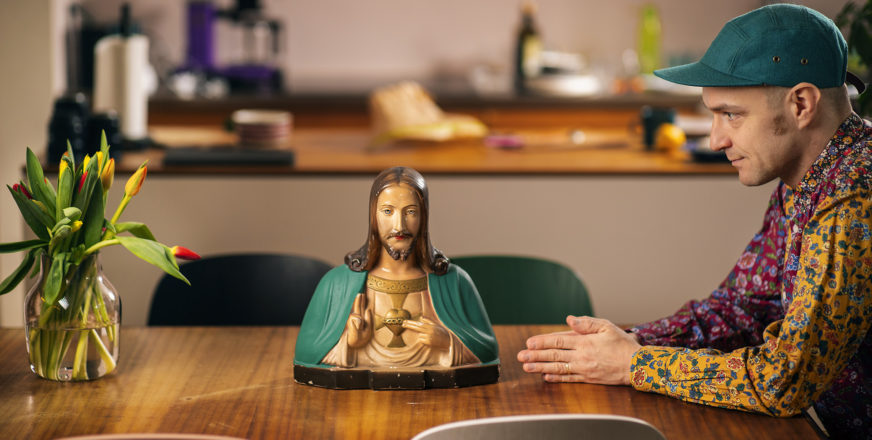 Jaakko Kämäräinen istuu sivuttain pöydän päässä ja katselee pöydällä olevaa Jeesus-patsasta