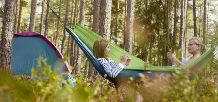 Kaksi ihmistä istuu riippukeinussa metsässä, taka-alalla teltta.