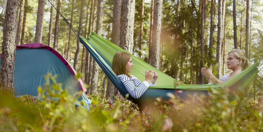 Kaksi ihmistä istuu riippukeinussa metsässä, taka-alalla teltta.