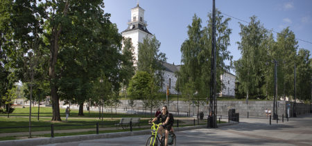 Kesämaisemassa Kuopion tuomiokirkko näkyy puiston ympäröimänä, etualalla kadulla pyöräilijä.