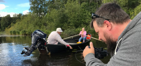 Kaksi ihmistä veneessä järvellä, kuvaaja videoimassa.