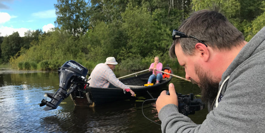 Kaksi ihmistä veneessä järvellä, kuvaaja videoimassa.