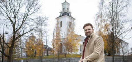 Jukka Voutilainen istuu kaiteella ja taustalla näkyy Kuopion tuomiokirkko sekä ruskan sävyttämiä puita