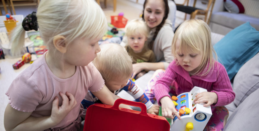 Pikkulapsia leikkimässä sohvalla, taustalla aikuinen. lapsi sylissään