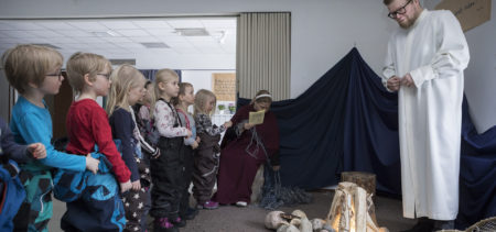 Seurakuntasalissa lapset kokoontuneena nuotioesityksen ääreen seuraamaan albaan pukeutunutta aikuista.