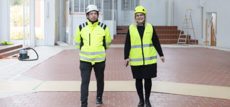 Urakoitsija ja kirkkoherra kävelevät hymyillen kameraa kohti Pyhän Johanneksen kirkon tyhjässä kirkkosalissa.