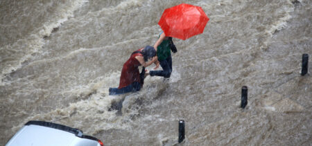 Sateessa ja tulvivalla kadulla käveleviä ihmisiä, punainen sateenvarro.