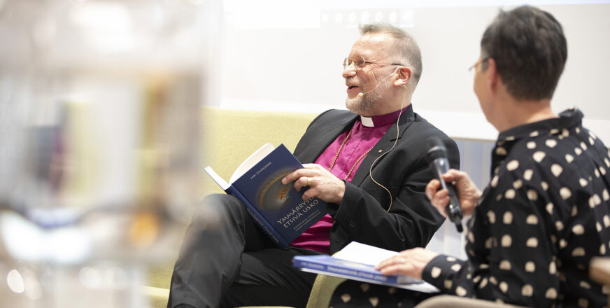 Jari Jolkkonen lehdistötilaisuudessa kirjan julkaisussa. Istuu sohvalla hymyillen avonainen kirja sylissä.