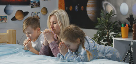 Kaksi lasta ja aikuinen polvistuneena rukoilemaan sängyn laidalle.