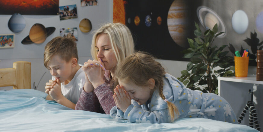 Kaksi lasta ja aikuinen polvistuneena rukoilemaan sängyn laidalle.