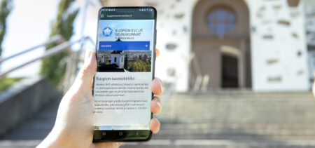 Kädessä älypuhelin, jonka näytöllä avoimena Tuomiokirkon verkkosivut, kirkko taustalla.