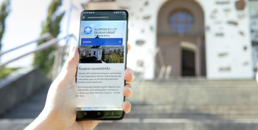 Kädessä älypuhelin, jonka näytöllä avoimena Tuomiokirkon verkkosivut, kirkko taustalla.