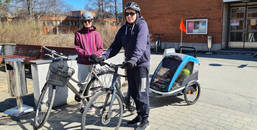 Kaksi aikuista pyörien ääressä, toisen pyörän perässä pyöräkärryssä kaksi lasta.