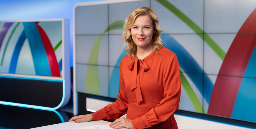 Uutisankkuri Piia Pasanen punaisessa asussa pöydän ääressä tv-studiossa