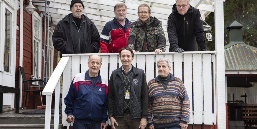 Seitsemän miestä ryhmäkuvassa: neljä mökin terassilla ja kolme terassin edessä