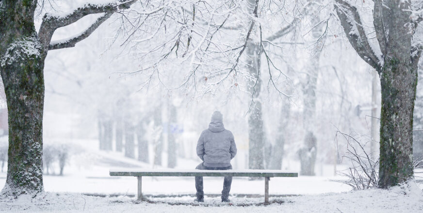 Harmaa talvinen maisema, ihminen istuu selin puistonpenkillä.