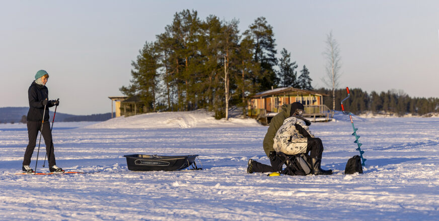 Hiihtäjä ja pilkkijä järven jäällä, taustalla pieni saari.