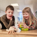 Kaksi lasta leikkii pehmoleluilla pöydän päällä ja nauraa.