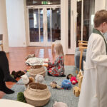 Äiti vauva sylissä, kaksi isompaa lasta leikkii kirkon leikkipaikalla.