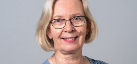 Kasvokuvassa hymyilevä Riitta Helle: vaaleat puolipitkät hiukset ja silmälasit.