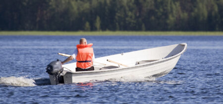 Järvellä pieni vene, jossa perämoottori ja yksi veneilijä pelastusliivit yllään.