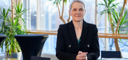 Ursula Schwab pöydän ääressä, katsoo kameraan ja hymyilee hieman. Taustalla viherkasveja ja isot ikkunat.