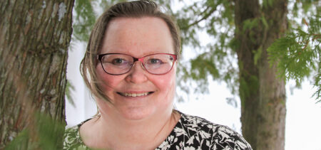 Marja-Leena Huttunen nojaa puunrunkoon, katsoo suoraan kameraan ja hymyilee. Taustalla puita ja vihreitä oksia.