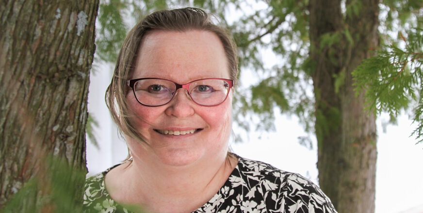 Marja-Leena Huttunen nojaa puunrunkoon, katsoo suoraan kameraan ja hymyilee. Taustalla puita ja vihreitä oksia.