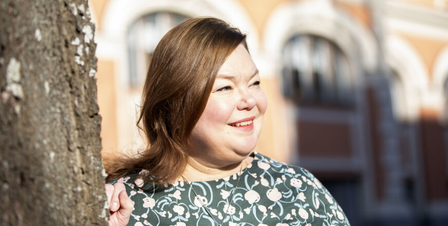 Minna Siikaniva nojaa puunrunkoon ja katsoo hymyillen ohi kameran, aurinko paistaa.