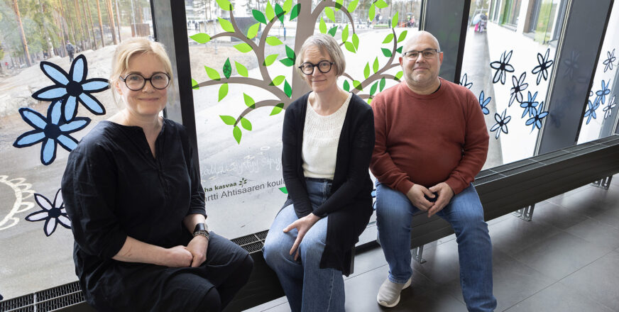 Kolme aikuista istuu ikkunan luona, ikkunassa askarreltu vihreälehtinen puu.