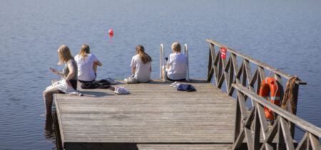 Nuorisoa istumassa laiturin nokassa selin. Aurinkoinen päivä ja sininen järvi.