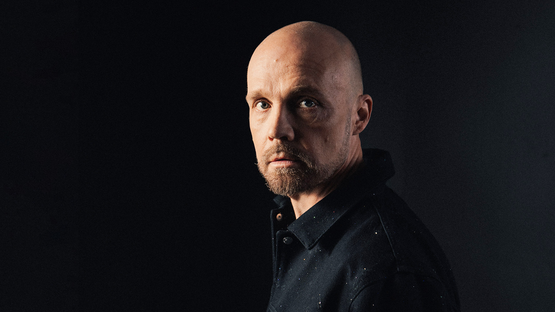 Juha Tapio seisoo sivuttain kasvot kääntyneenä kohti kameraa. Tausta on täysin musta.