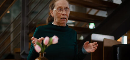 Kati Outinen puhuu ja levittelee käsiään, taustalla portaat ja etualalla kukkia maljakossa