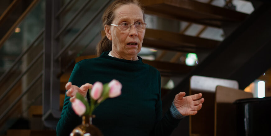 Kati Outinen puhuu ja levittelee käsiään, taustalla portaat ja etualalla kukkia maljakossa